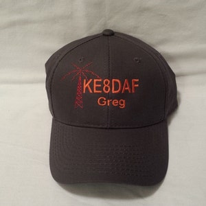 May include: A dark gray baseball cap featuring an embroidered red radio tower icon on the left side. To the right of the icon, the text KE8DAF is stitched in orange, with the name Greg embroidered in orange directly underneath the call sign.