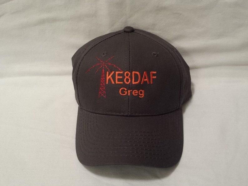 May include: A dark gray baseball cap featuring an embroidered red radio tower icon on the left side. To the right of the icon, the text KE8DAF is stitched in orange, with the name Greg embroidered in orange directly underneath the call sign.