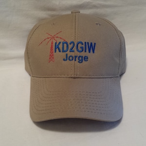 May include: A tan baseball cap featuring an embroidered red radio tower icon next to the blue text KD2GIW and the name Jorge in blue embroidery. The cap has a structured crown, a curved brim with stitched detailing, and a button on top.