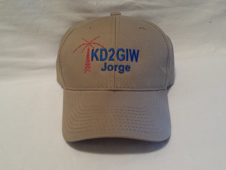 May include: A tan baseball cap featuring an embroidered red radio tower icon next to the blue text KD2GIW and the name Jorge in blue embroidery. The cap has a structured crown, a curved brim with stitched detailing, and a button on top.