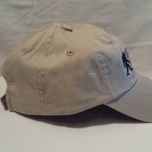 May include: A beige baseball cap with a black and red embroidered design of two figures walking. The cap has a curved brim and a mesh back.