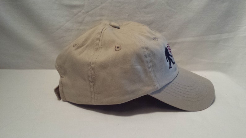 May include: A beige baseball cap with a black and red embroidered design of two figures walking. The cap has a curved brim and a mesh back.