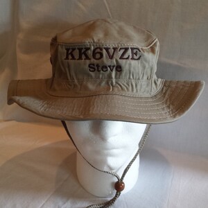 May include: Tan bucket hat with a wide brim and a chin strap. The hat features embroidered text in a darker brown color that reads "KK6VZE" above "Steve". The hat is displayed on a white mannequin head.