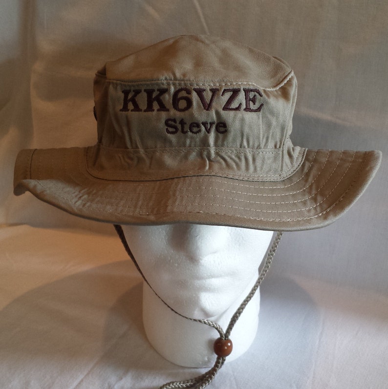 May include: Tan bucket hat with a wide brim and a chin strap. The hat features embroidered text in a darker brown color that reads "KK6VZE" above "Steve". The hat is displayed on a white mannequin head.