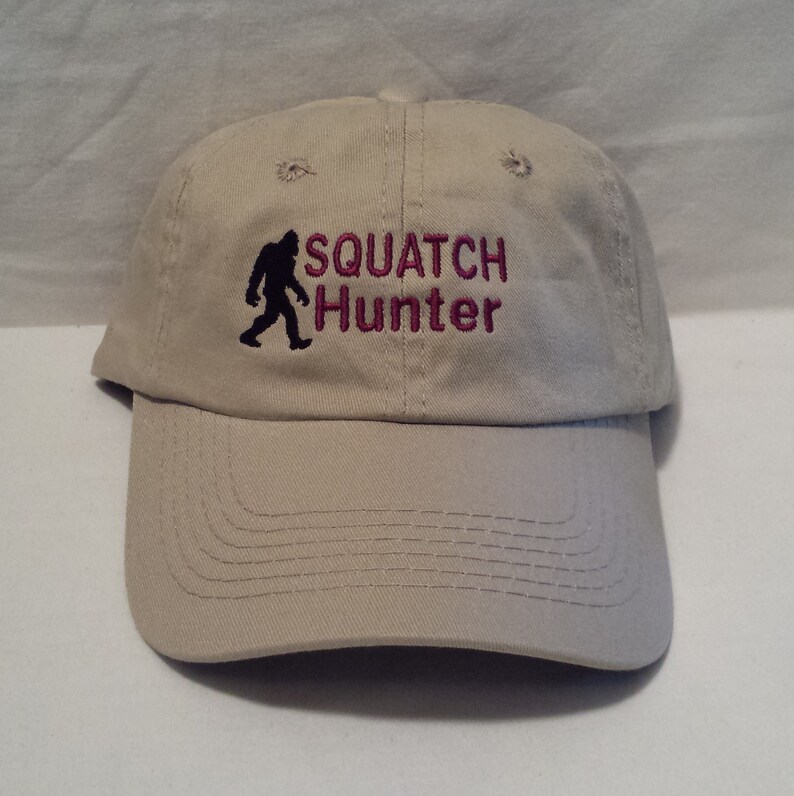 May include: A beige baseball cap with a black silhouette of a Bigfoot and the text "SQUATCH Hunter" in pink embroidery.