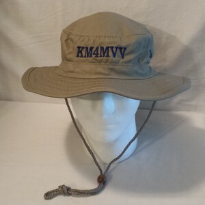 May include: Tan bucket hat with a wide brim and a chin strap. The hat features the embroidered text "KM4MVV" in blue on the front. A small metal button is on the side of the hat.