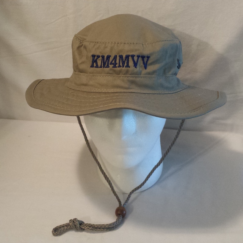 May include: Tan bucket hat with a wide brim and a chin strap. The hat features the embroidered text "KM4MVV" in blue on the front. A small metal button is on the side of the hat.