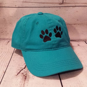 May include: A teal blue baseball cap with two black paw prints embroidered on the front.