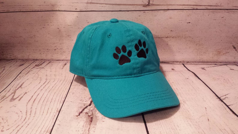 May include: A teal blue baseball cap with two black paw prints embroidered on the front.