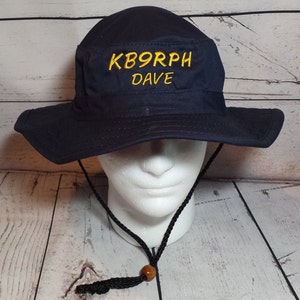 May include: Navy blue bucket hat with a wide brim and chin strap. The hat features the embroidered text "KB9RPH DAVE" in yellow. The hat is displayed on a white mannequin head.