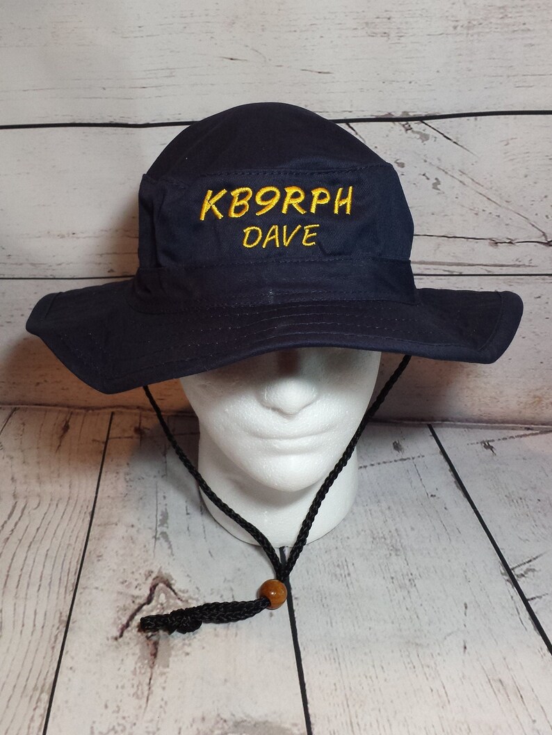 May include: Navy blue bucket hat with a wide brim and chin strap. The hat features the embroidered text "KB9RPH DAVE" in yellow. The hat is displayed on a white mannequin head.