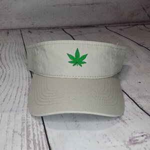 May include: A beige visor with a green marijuana leaf embroidered on the front.