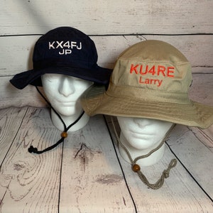 May include: Two bucket hats on white mannequin heads. One is navy blue with white embroidered text "KX4FJ JP". The other is tan with red embroidered text "KU4RE Larry". Both hats have chin straps with wooden beads.