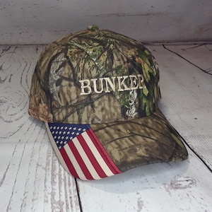 May include: A camouflage baseball cap with a white embroidered "BUNKER" on the front and an American flag patch on the side.