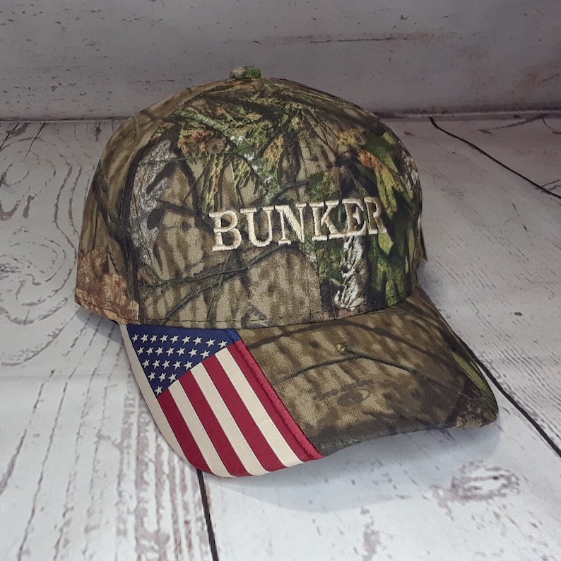 May include: A camouflage baseball cap with a white embroidered "BUNKER" on the front and an American flag patch on the side.