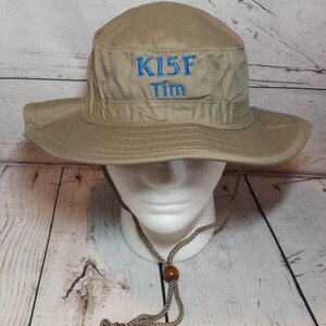 May include: A tan-colored bucket hat with a wide brim and a chin strap. The hat features the embroidered text "KI5F Tim" in blue thread on the front. The hat is displayed on a white mannequin head.