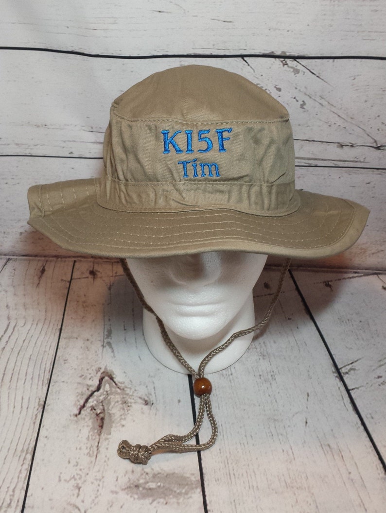 May include: A tan-colored bucket hat with a wide brim and a chin strap. The hat features the embroidered text "KI5F Tim" in blue thread on the front. The hat is displayed on a white mannequin head.