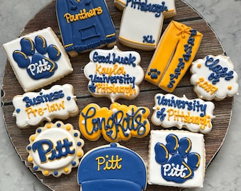 Puede incluir: Un conjunto de 12 galletas de azúcar decoradas con glaseado azul y dorado. Las galletas presentan el logotipo de la Universidad de Pittsburgh, las palabras "Pitt", "Panthers", "Congrats", "University of Pittsburgh", "Class of 2023", "Good Luck Kayla University of Pittsburgh", "Business Pre-Law", y una gorra de graduación.