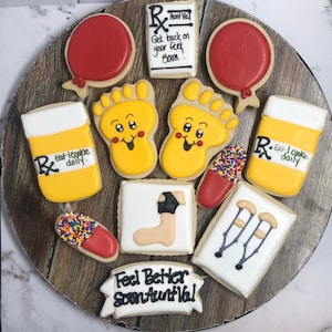 Könnte beinhalten: Ein Set dekorierter Zuckerkekse mit einem Gute-Besserung-Motiv. Die Kekse sind mit Bildern von Füßen, Krücken, Pillen und einer Rezeptflasche verziert. Die Kekse sind auf einem Holzteller angeordnet. Der Text auf den Keksen lautet "Get back on your feet soon", "Eat I cookie daily", und "Feel Better Soon Aunt Val".