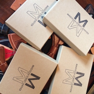 May include: Five brown cardboard boxes with the text "Madison Street Leather" and a stylised "M" logo. The boxes are arranged in a pyramid shape with the largest box at the bottom and the smallest box at the top. The boxes are surrounded by various brown leather bow ties.