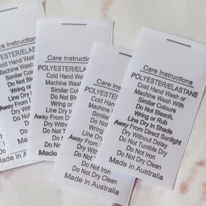May include: Six white care instruction labels with black text. The labels are for polyester/elastane fabric and include instructions for washing, drying, and ironing. The labels are made in Australia. A black and white image of a sewing machine with the text "RUBYJAM FABRIC" is in the background.