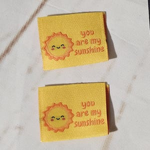 Puede incluir: Etiquetas de tela amarillas con el texto "You are my sunshine" y un gráfico de sol sonriente. Algunas etiquetas están sueltas, otras en una caja negra. También hay una tarjeta blanca con un gráfico de máquina de coser.