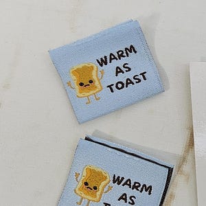 Puede incluir: Dos etiquetas de tela azul claro con el texto "WARM AS TOAST" y una ilustración de tostada de dibujos animados. También es visible una pegatina en blanco y negro con un diseño de máquina de coser y el texto "RUBYJAM FABRIC".