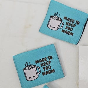 Puede incluir: Varias etiquetas de tela turquesa con una taza de chocolate caliente de dibujos animados y el texto "MADE TO KEEP YOU WARM". Un logotipo de RubyJam Fabric en blanco y negro muestra una silueta de máquina de coser con un corazón.