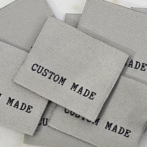 Puede incluir: Un conjunto de etiquetas de tela gris con la inscripción "CUSTOM MADE" en negro. Las etiquetas rectangulares tienen una textura. Están dispuestas sobre una superficie de madera clara.