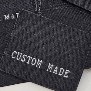 Puede incluir: Un conjunto de etiquetas cuadradas de tela negra con la inscripción "CUSTOM MADE" bordada en blanco. Las etiquetas, hechas de material tejido, están dispuestas sobre una superficie clara.