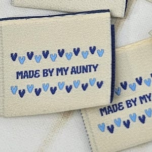 Puede incluir: Varias etiquetas de tela blanquecina con patrones de corazones azules y el texto "MADE BY MY AUNTY" en azul. Las etiquetas tienen un borde azul oscuro y están dispuestas sobre una superficie clara.