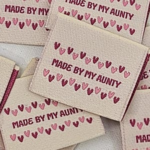 Puede incluir: Un conjunto de etiquetas de tela beige con el texto "MADE BY MY AUNTY" en rosa, enmarcadas por corazones rosas. Las etiquetas son rectangulares y tienen un borde doblado. El texto es en rosa.