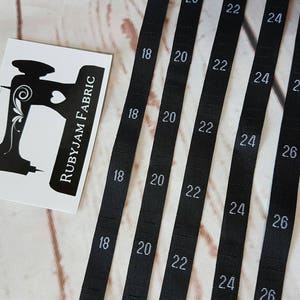 Puede incluir: Cinco etiquetas de cinta de tela negra con texto blanco. Las etiquetas están numeradas 18, 20, 22, 24 y 26.