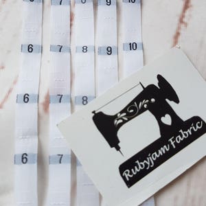 Puede incluir: Cinco cintas métricas de tela blanca con los números 6, 7, 8, 9 y 10 impresos en ellas. Una pegatina blanca con una ilustración de máquina de coser negra y el texto "Rubyjam Fabric" está encima de las cintas.