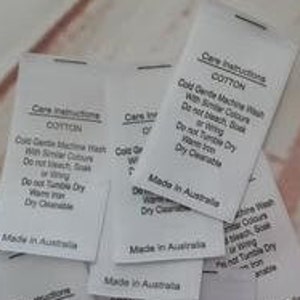 Puede incluir: Una pila de etiquetas blancas de instrucciones de cuidado para telas de algodón. Las etiquetas tienen texto negro que dice "Care Instructions Cotton Cold Gentle Machine Wash With Similar Colours Do Not Bleach, Soak or Wring Do Not Tumble Dry Warm Iron Dry Cleanable Made in Australia". En el fondo, una silueta en blanco y negro de una máquina de coser con el texto "RUBYJAM FABRIC".