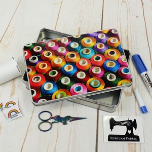 Puede incluir: Una caja de metal plateada con un estampado colorido de carretes de hilo. La caja está abierta y la tapa está apoyada en la parte superior. La caja está rodeada de varios artículos de costura, incluyendo tijeras, un rollo de cinta blanca, dos pequeñas pegatinas de arcoíris y dos marcadores blancos.