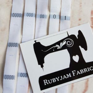 Puede incluir: Cinta de tela blanca con números impresos. La cinta está etiquetada con "RUBYJAM FABRIC" y presenta una silueta negra de una máquina de coser con un corazón.
