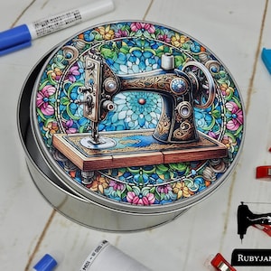 Puede incluir: Una lata redonda de metal con un diseño de vitral colorido que presenta una máquina de coser vintage. El diseño incluye elementos florales y un fondo azul y verde. La lata es plateada y tiene un borde ligeramente elevado.