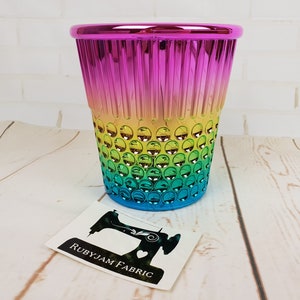 Puede incluir: Un dedal de metal de color arcoíris con una superficie texturizada. El dedal está sobre una superficie de madera blanca. El texto "RubyJam Fabric" es visible en una pegatina en primer plano.