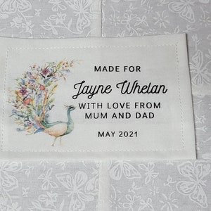 Könnte beinhalten: Ein weißes Stofflabel mit einem Pfau und einem Blumenmuster. Das Etikett lautet "Made for Jayne Whelan with love from Mum and Dad May 2021".