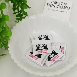 Puede incluir: Un conjunto de 10 etiquetas tejidas blancas con corazones negros en forma de mano y el texto "hecho a mano con amor" en rosa.