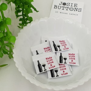 Puede incluir: Etiquetas tejidas blancas con el texto "Made with love & wine" en rojo y negro, con una ilustración de una botella y una copa de vino. Las etiquetas están en un plato blanco festoneado. La etiqueta superior dice "JOSIE BUTTONS 10 Woven Labels."