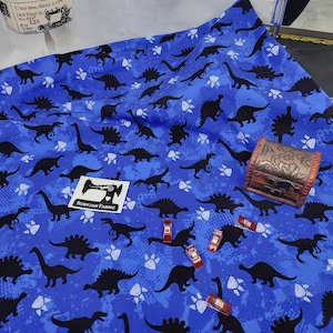 May include: Blue fabric with a repeating pattern of black dinosaurs and white paw prints. The fabric has a distressed look and a tag that says "Rubyjam Fabric".