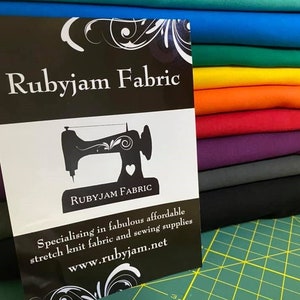 May include: A black and white sign with the text "Rubyjam Fabric" and a sewing machine graphic. The sign is in front of a stack of colorful fabric swatches.