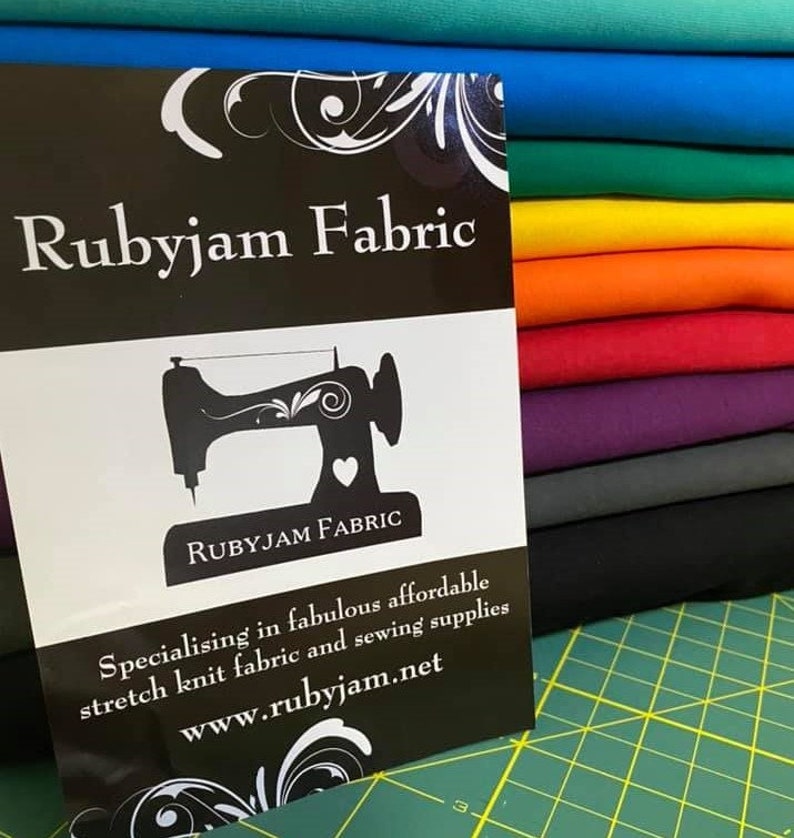 May include: A black and white sign with the text "Rubyjam Fabric" and a sewing machine graphic. The sign is in front of a stack of colorful fabric swatches.