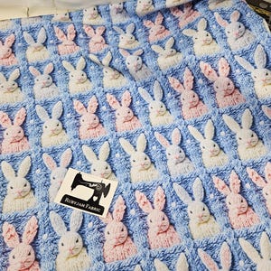 Puede incluir: Tejido de punto azul con un patrón repetido de caras de conejos en blanco y rosa. El tejido tiene un aspecto texturizado, con cada cara de conejo en relieve. Se ve una pequeña etiqueta con el logotipo de una máquina de coser.