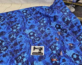 May include: Blue fabric with a soccer ball pattern. The fabric features a repeating design of soccer balls in varying shades of blue against a textured background. The fabric is decorated with several red and silver clips and a label that says "RUBYJAM FABRIC".