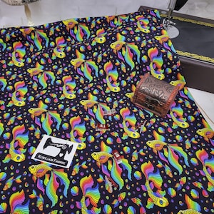 Puede incluir: Tela negra con un patrón repetido de peces dorados arcoíris y conchas marinas. La tela está impresa con el texto "Rubyjam Fabric".