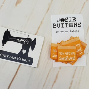 Puede incluir: Una tarjeta blanca con el texto "JOSIE BUTTONS" y "10 Woven Labels" junto a una pila de etiquetas naranjas que dicen "You are my Sunshine". También hay una tarjeta en blanco y negro con un gráfico de máquina de coser y el texto "RUBYJAM FABRIC".