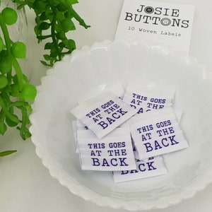 Puede incluir: Diez etiquetas tejidas blancas con el texto "This goes at the back" impreso en morado. Las etiquetas están en un tazón blanco.